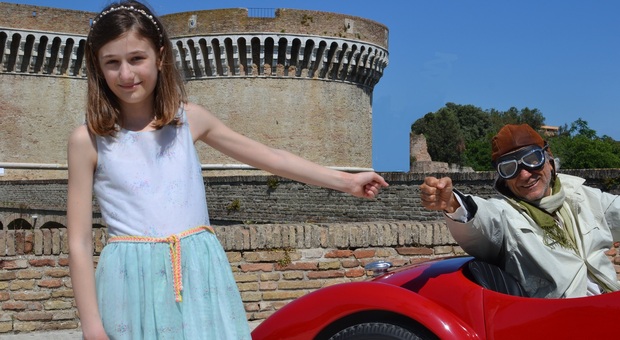 Elena Schiavoni nei panni della bambina, protagonista del ritrovamento delle bobine con le corse, mentre saluta il passaggio di un’auto storica davanti alla Rocca roveresca di Senigallia